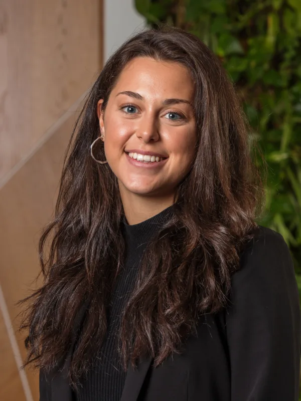 Person standing indoors wearing a black blazer and dark top, with long dark hair over the shoulders. The background features light wood panelling and a vertical green plant wall.