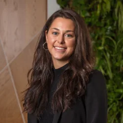 Person standing indoors wearing a black blazer and dark top, with long dark hair over the shoulders. The background features light wood panelling and a vertical green plant wall.