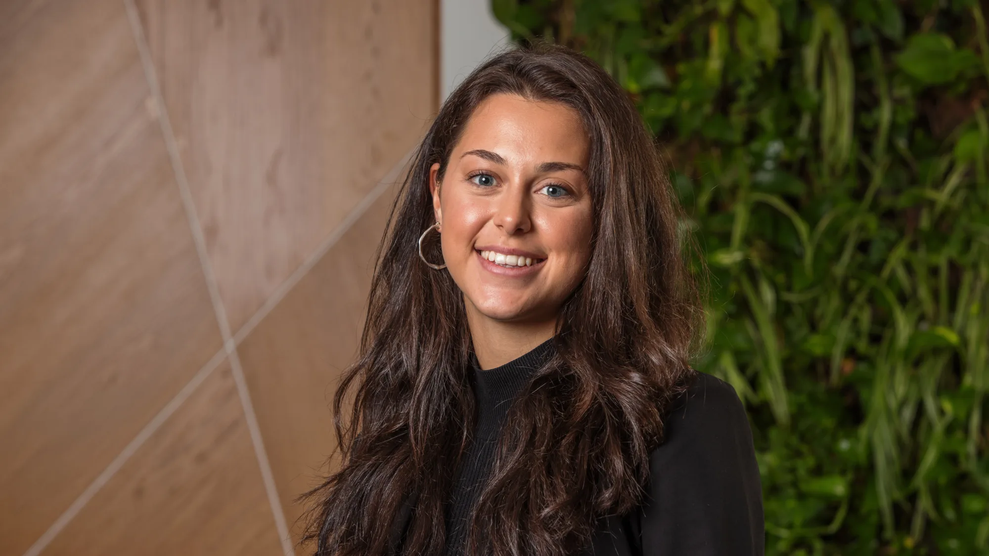 Person standing indoors wearing a black blazer and dark top, with long dark hair over the shoulders. The background features light wood panelling and a vertical green plant wall.