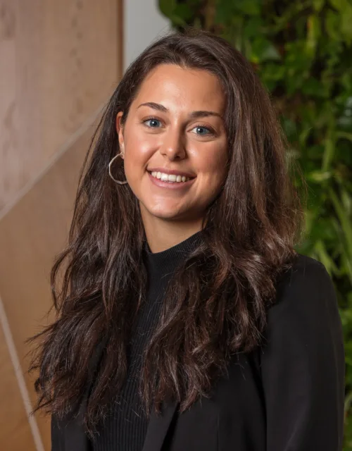Person standing indoors wearing a black blazer and dark top, with long dark hair over the shoulders. The background features light wood panelling and a vertical green plant wall.