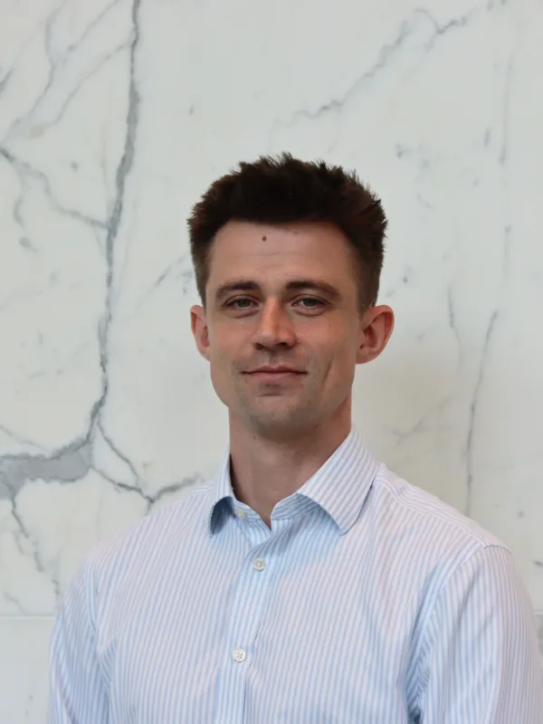 Person in a light blue and white striped button-down shirt standing indoors against a white marble wall with grey veining. The image is a centered head-and-shoulders portrait with no visible text or additional objects.