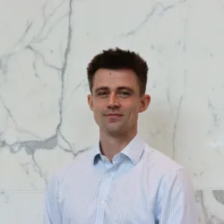 Person in a light blue and white striped button-down shirt standing indoors against a white marble wall with grey veining. The image is a centered head-and-shoulders portrait with no visible text or additional objects.