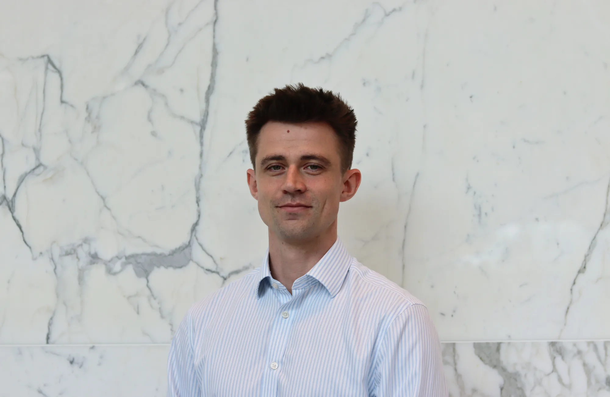 Person in a light blue and white striped button-down shirt standing indoors against a white marble wall with grey veining. The image is a centered head-and-shoulders portrait with no visible text or additional objects.