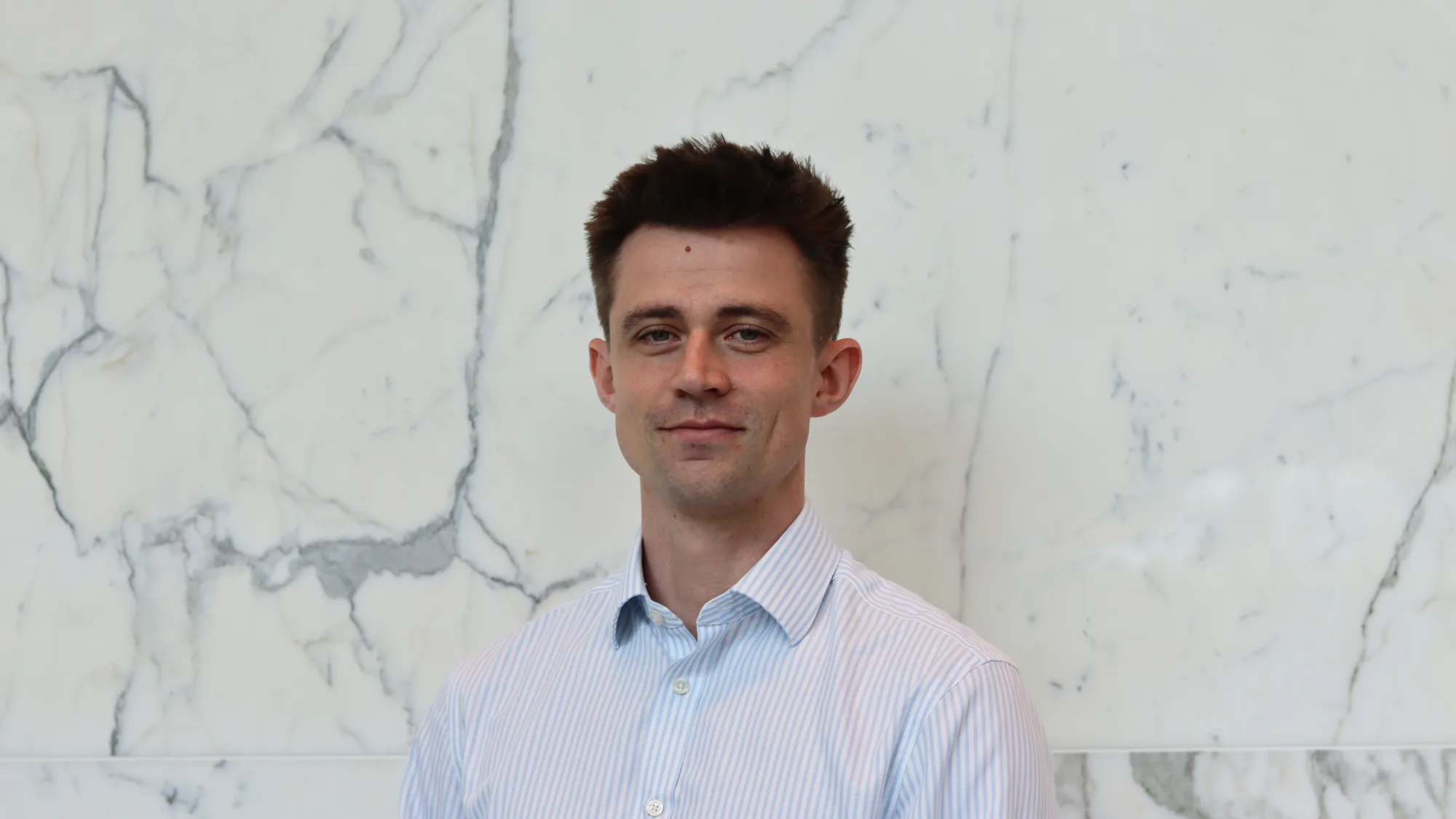 Person in a light blue and white striped button-down shirt standing indoors against a white marble wall with grey veining. The image is a centered head-and-shoulders portrait with no visible text or additional objects.