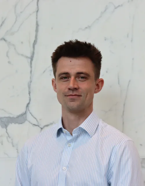 Person in a light blue and white striped button-down shirt standing indoors against a white marble wall with grey veining. The image is a centered head-and-shoulders portrait with no visible text or additional objects.