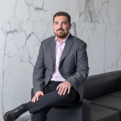 Curtis Gubb sitting in a grey blazer on the arm of a black sofa next to a marble tiled wall