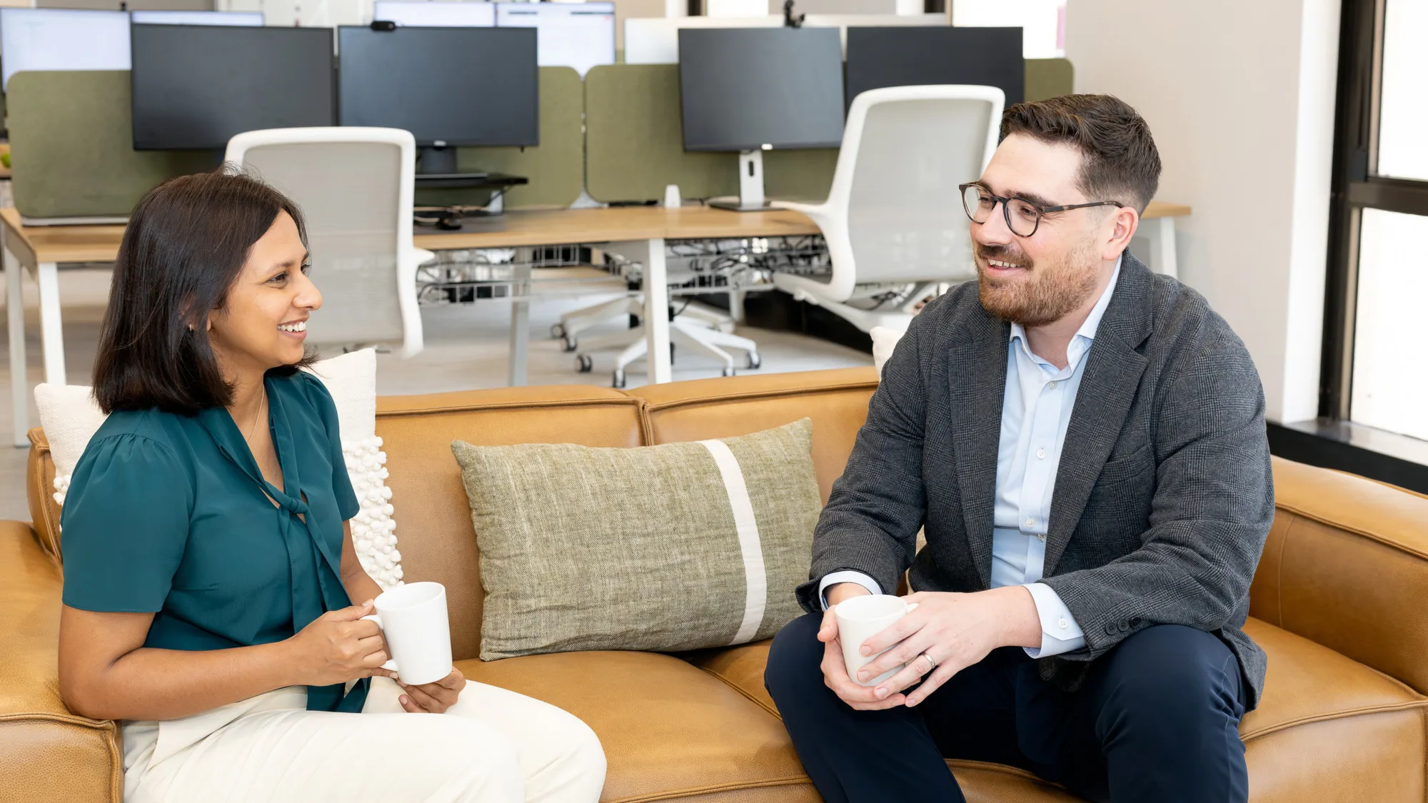 Two people sit on a tan sofa in an open-plan office, holding mugs while talking. A table with books is in the foreground, and desks with monitors and chairs are visible behind them.