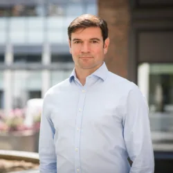 Head and sholders shot of Dan Bennett in blue shirt and black trousers with buildings in back ground