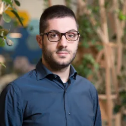 Danilo Cava in London office reception wearing a dark blue shirt with wooden panelling and plants in background