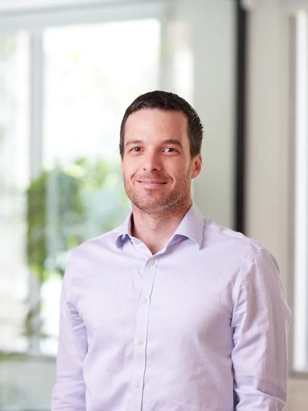 David in a pale open collared shirt in front of a glazed meeting room smiling to camera