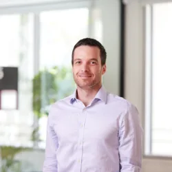 David in a pale open collared shirt in front of a glazed meeting room smiling to camera