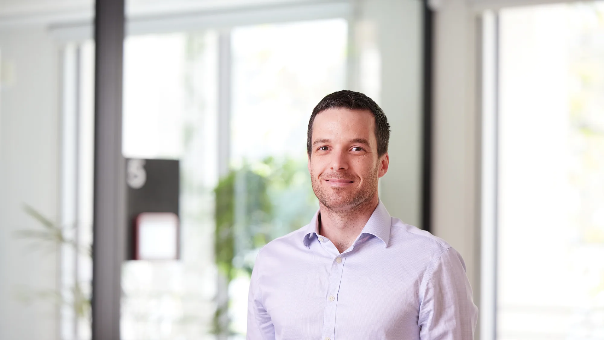 David in a pale open collared shirt in front of a glazed meeting room smiling to camera