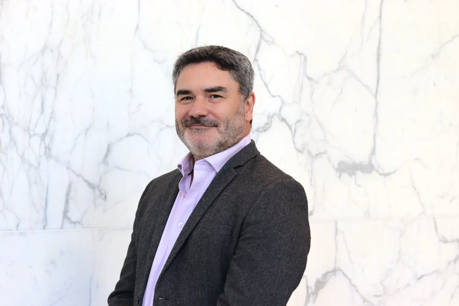 Head and sholder shot of Drew against a white marble background wearing a pale pink shirt and grey blazer