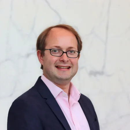Person with short hair wearing a dark blazer and light pink collared shirt stands in front of a white marble wall with gray veining.