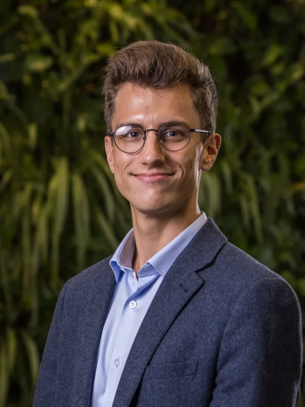 Laszlo in a pale blue open collar shirt and suit jacket in front of a green living wall smiling to camera