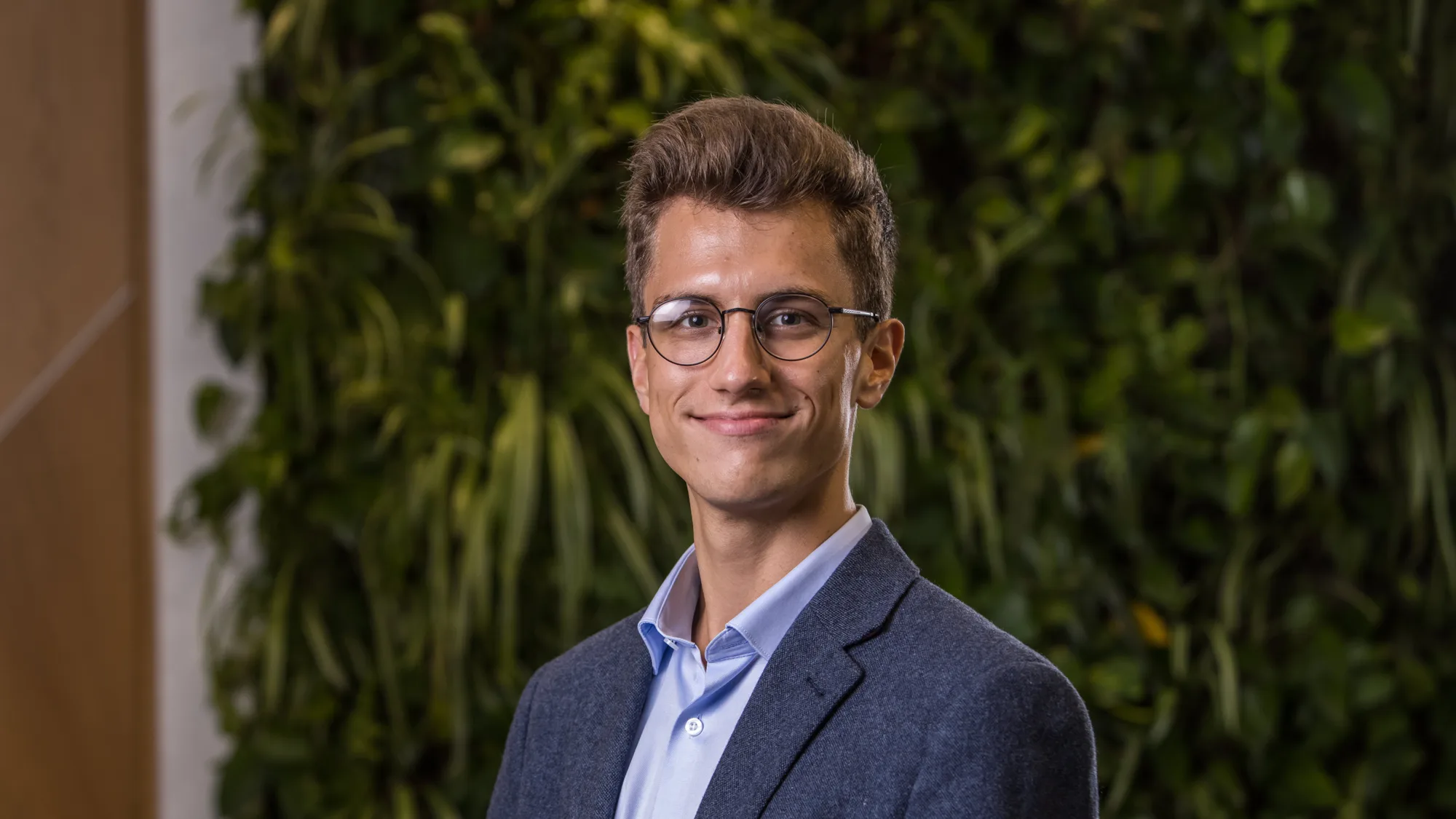 Laszlo in a pale blue open collar shirt and suit jacket in front of a green living wall smiling to camera