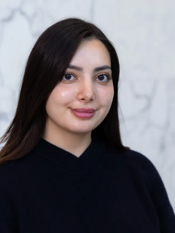 A person with straight dark hair wearing a dark V‑neck sweater is shown from the shoulders up, standing against a light grey marble wall with visible veining patterns in the background.