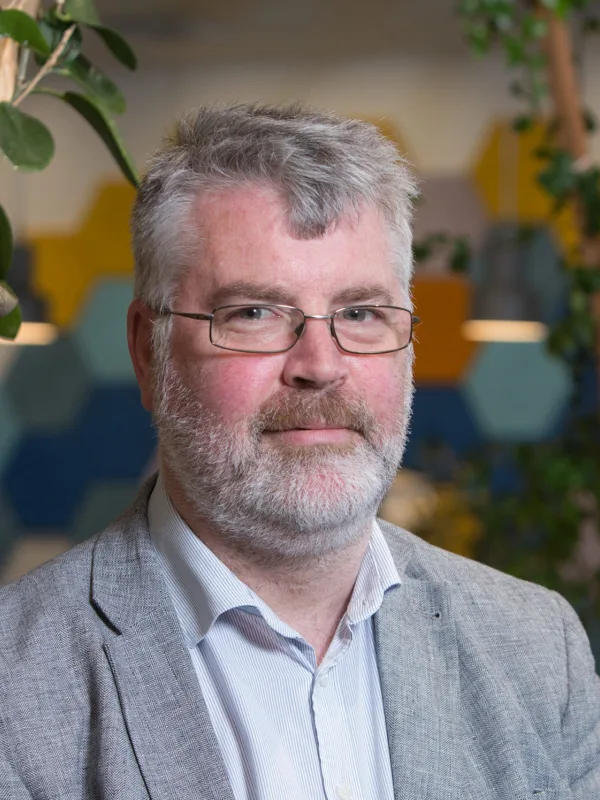 Head and shoulders image of Peter Owens in an office stood in front of shelves covered with green plants wearing a grey suit jacket and a plain blue shirt