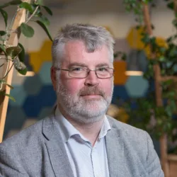 Head and shoulders image of Peter Owens in an office stood in front of shelves covered with green plants wearing a grey suit jacket and a plain blue shirt