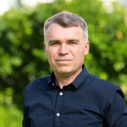 Head and sholders shot of Rasti wearing a dark blue shirt against green foliage
