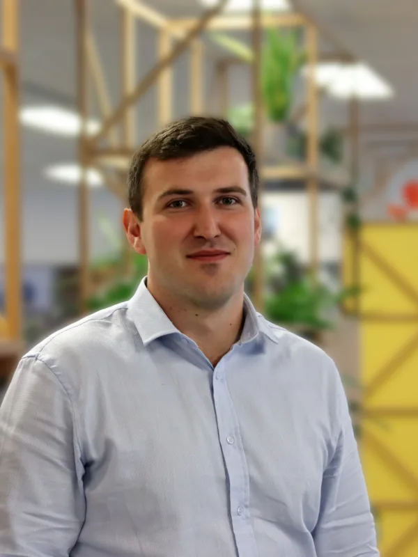 Person standing indoors in a modern office, wearing a light blue button-down shirt. Wooden shelving with plants and a yellow partition appear behind, with desks and other people in the background.