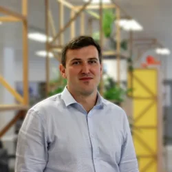 Person standing indoors in a modern office, wearing a light blue button-down shirt. Wooden shelving with plants and a yellow partition appear behind, with desks and other people in the background.