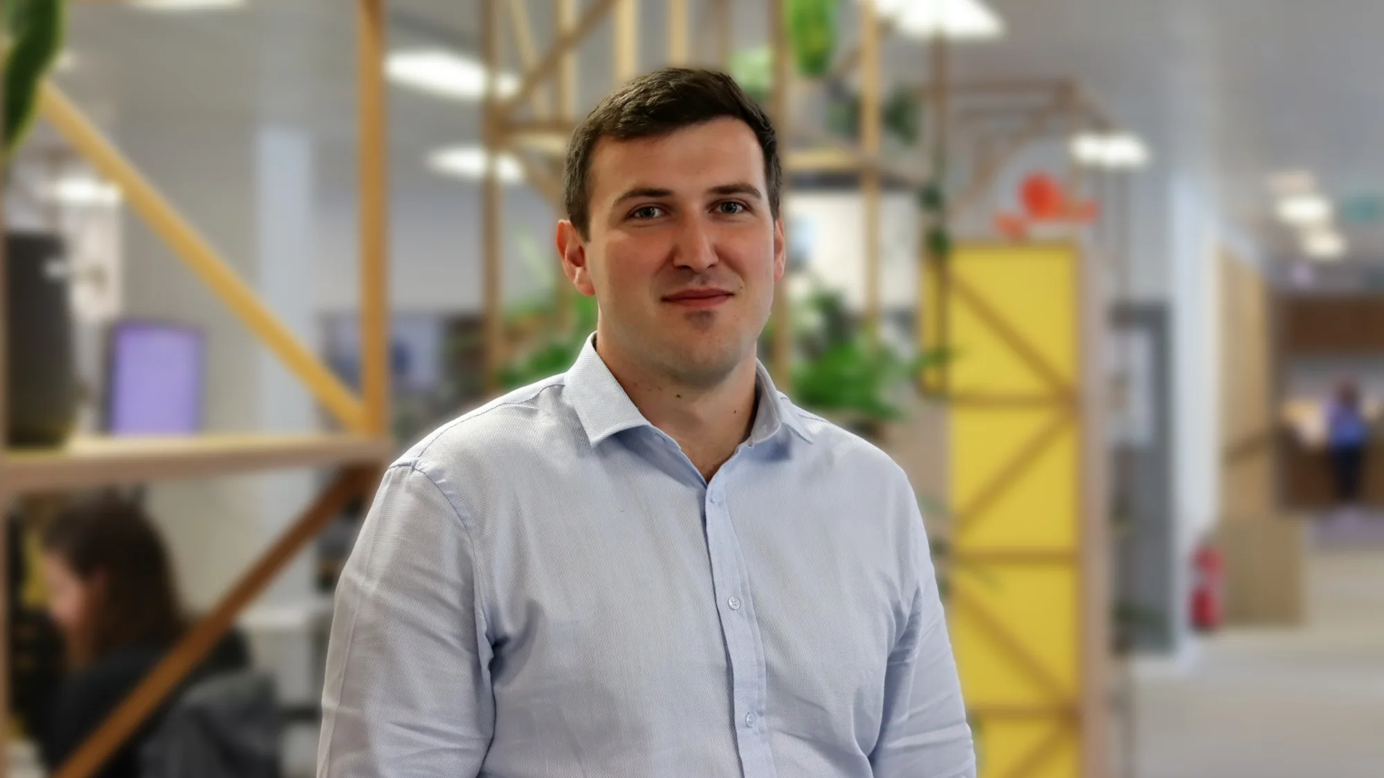Person standing indoors in a modern office, wearing a light blue button-down shirt. Wooden shelving with plants and a yellow partition appear behind, with desks and other people in the background.