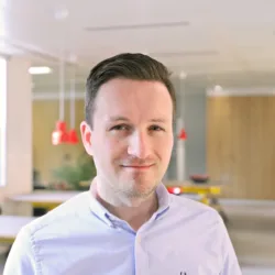 Man with short dark hair wearing a light blue button-up shirt stands in a modern office. Background includes people at tables with blurred faces, wooden elements, red pendant lights, and large windows.