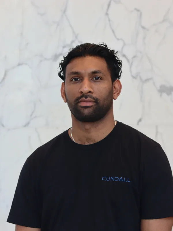 A person with dark curly hair stands against a light marble wall, wearing a black T‑shirt with the word ‘Cundall’ printed in blue on the chest. Upper body is centered in the frame.