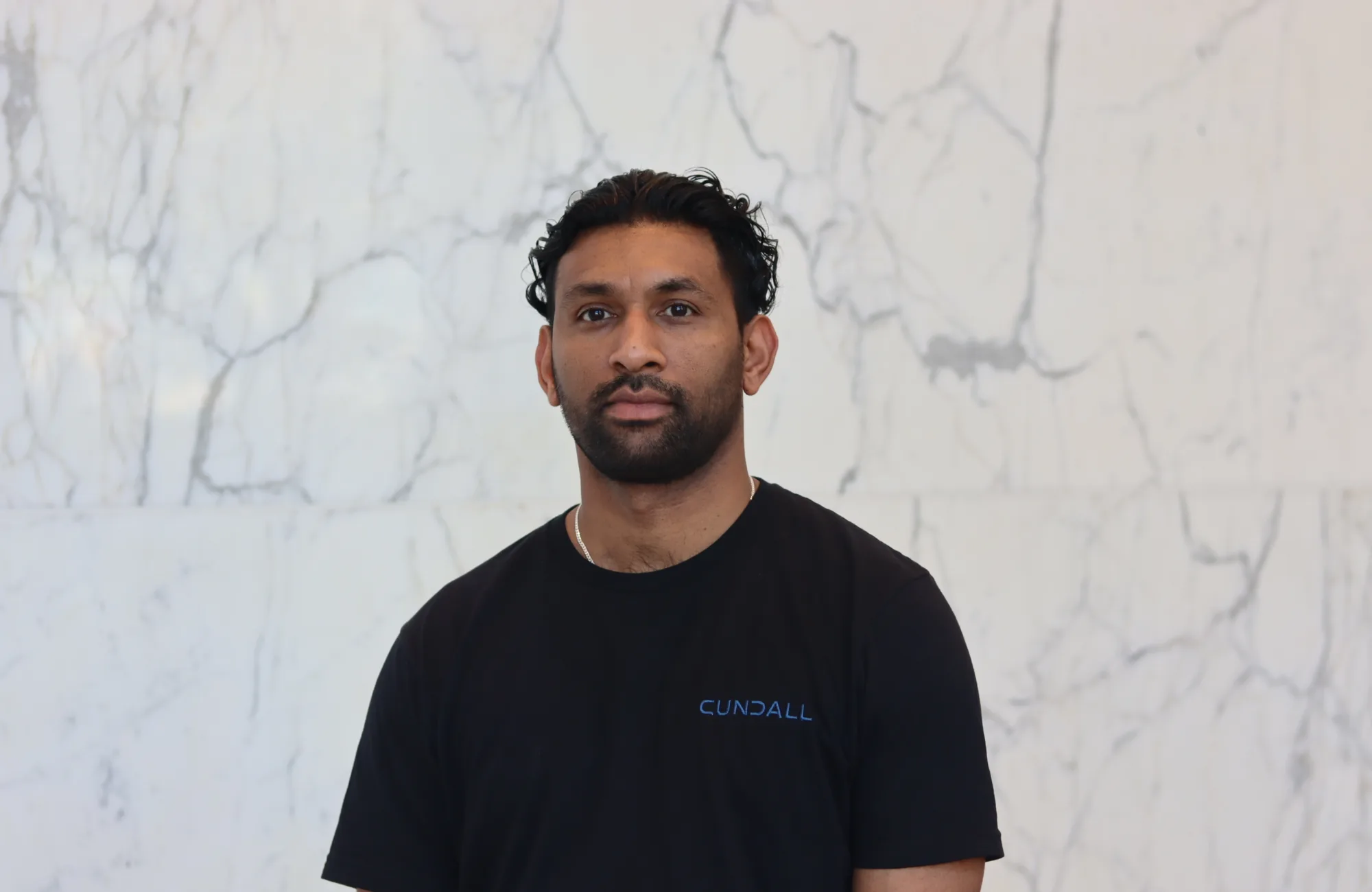 A person with dark curly hair stands against a light marble wall, wearing a black T‑shirt with the word ‘Cundall’ printed in blue on the chest. Upper body is centered in the frame.
