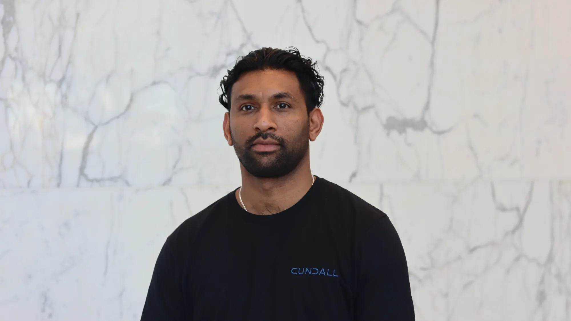 A person with dark curly hair stands against a light marble wall, wearing a black T‑shirt with the word ‘Cundall’ printed in blue on the chest. Upper body is centered in the frame.