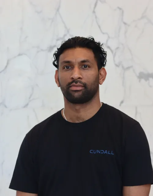 A person with dark curly hair stands against a light marble wall, wearing a black T‑shirt with the word ‘Cundall’ printed in blue on the chest. Upper body is centered in the frame.