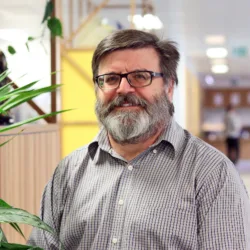 Wayne in an open collar shirt stood in an open plan office