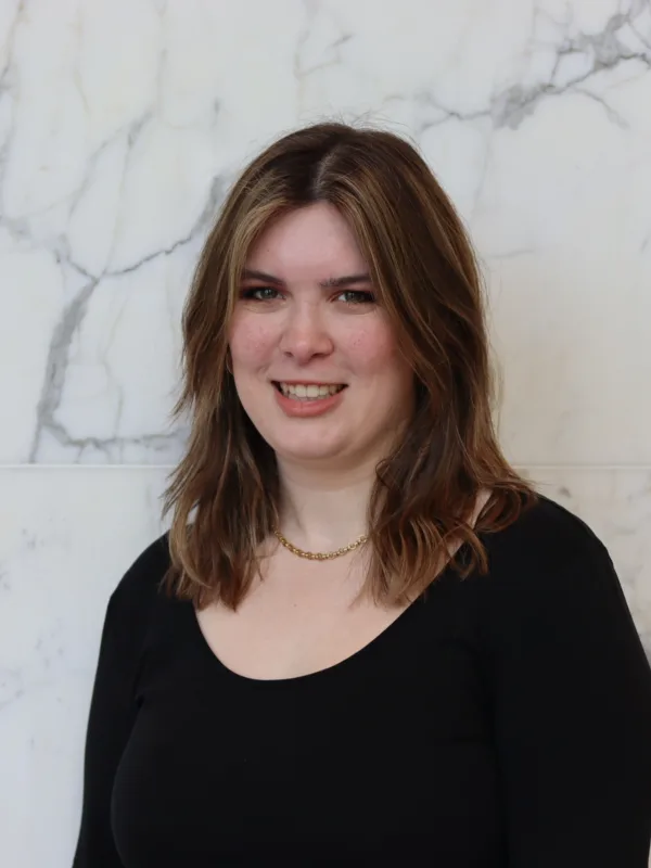 Portrait photograph of a person with shoulder-length brown hair, wearing a black long-sleeve top and a short gold necklace, standing against a white marble wall with grey veining. No visible text.