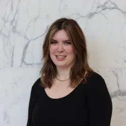 Portrait photograph of a person with shoulder-length brown hair, wearing a black long-sleeve top and a short gold necklace, standing against a white marble wall with grey veining. No visible text.