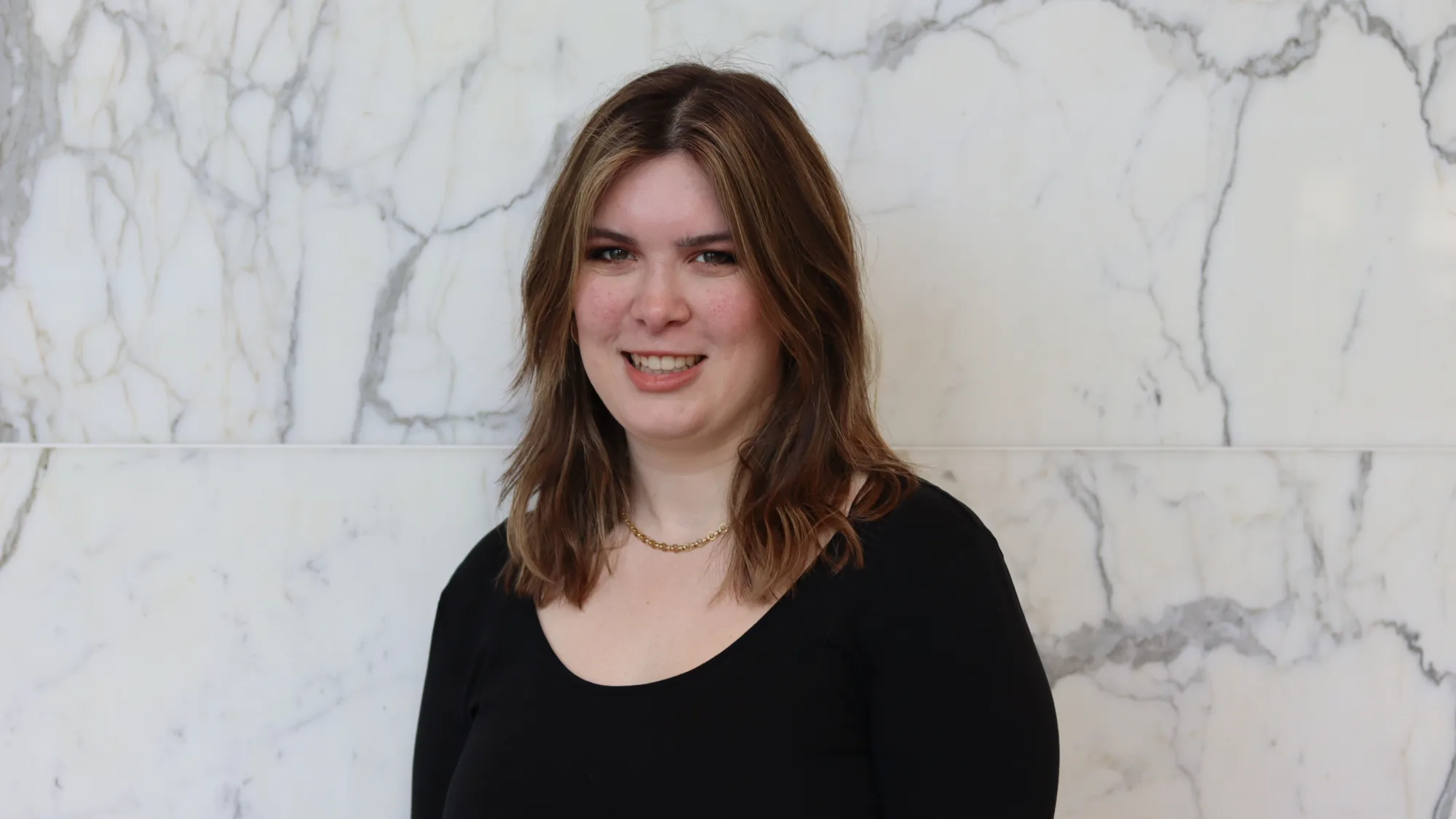 Portrait photograph of a person with shoulder-length brown hair, wearing a black long-sleeve top and a short gold necklace, standing against a white marble wall with grey veining. No visible text.
