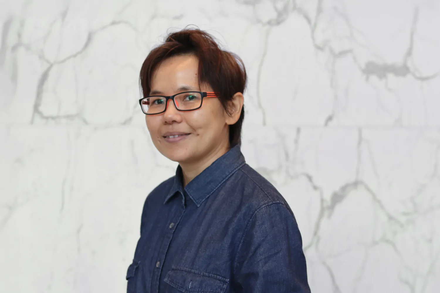 A person wearing glasses and a dark blue denim shirt stands in front of a white marble wall with gray veining. No text or additional objects are present.