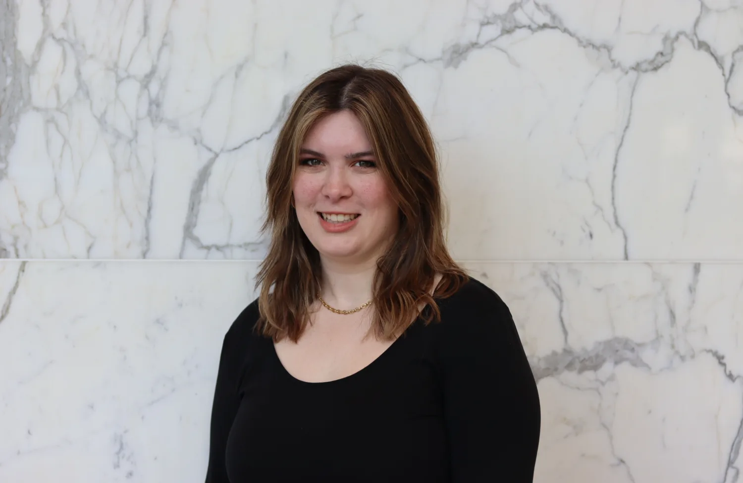 Portrait photograph of a person with shoulder-length brown hair, wearing a black long-sleeve top and a short gold necklace, standing against a white marble wall with grey veining. No visible text.