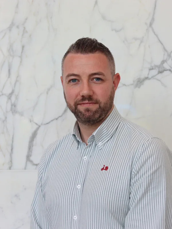 Ciaran in a pale open collar shirt smiling to camera in front of a marble texture wall