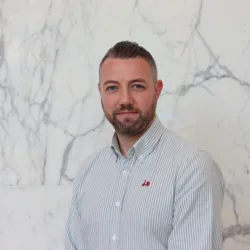 Ciaran in a pale open collar shirt smiling to camera in front of a marble texture wall