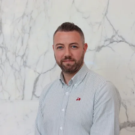 Ciaran in a pale open collar shirt smiling to camera in front of a marble texture wall