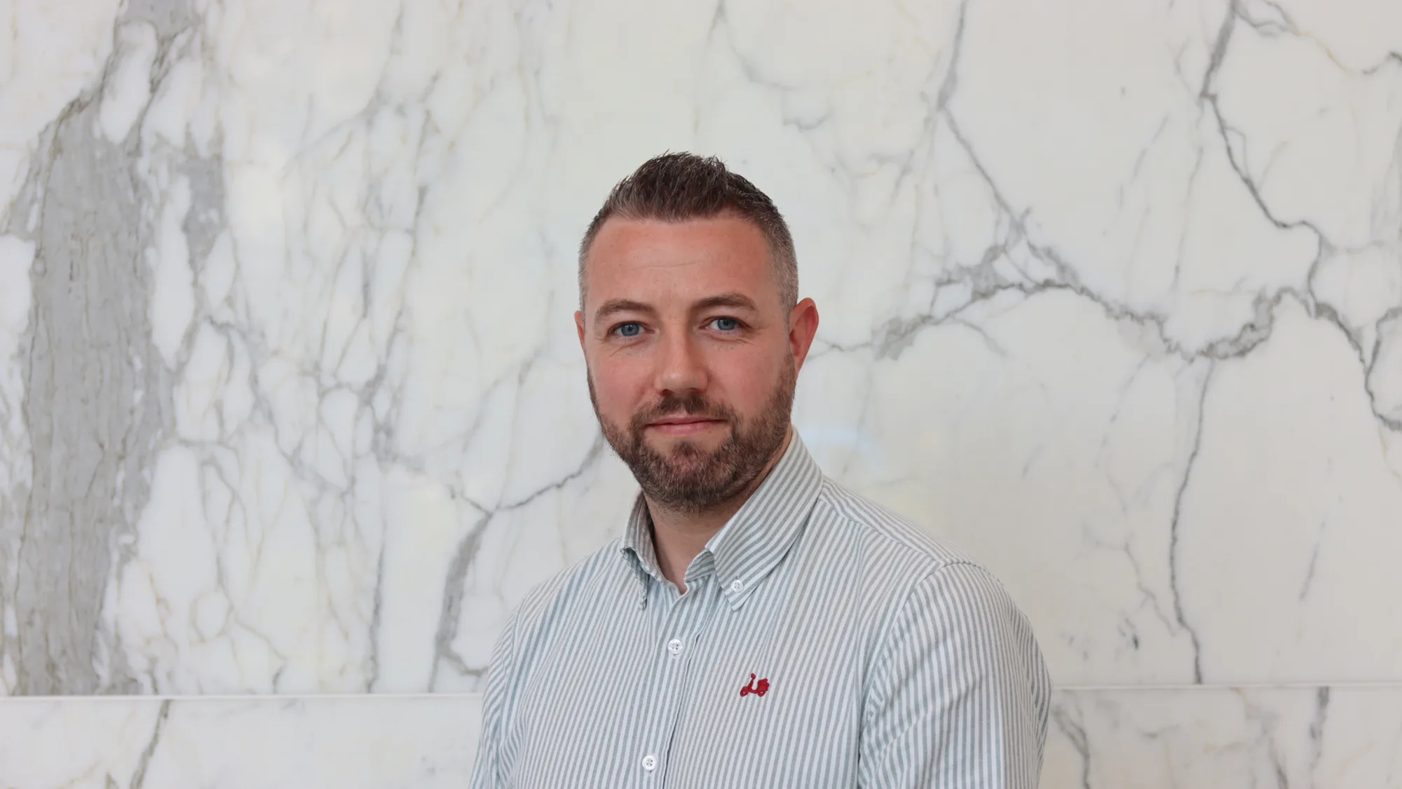 Ciaran in a pale open collar shirt smiling to camera in front of a marble texture wall