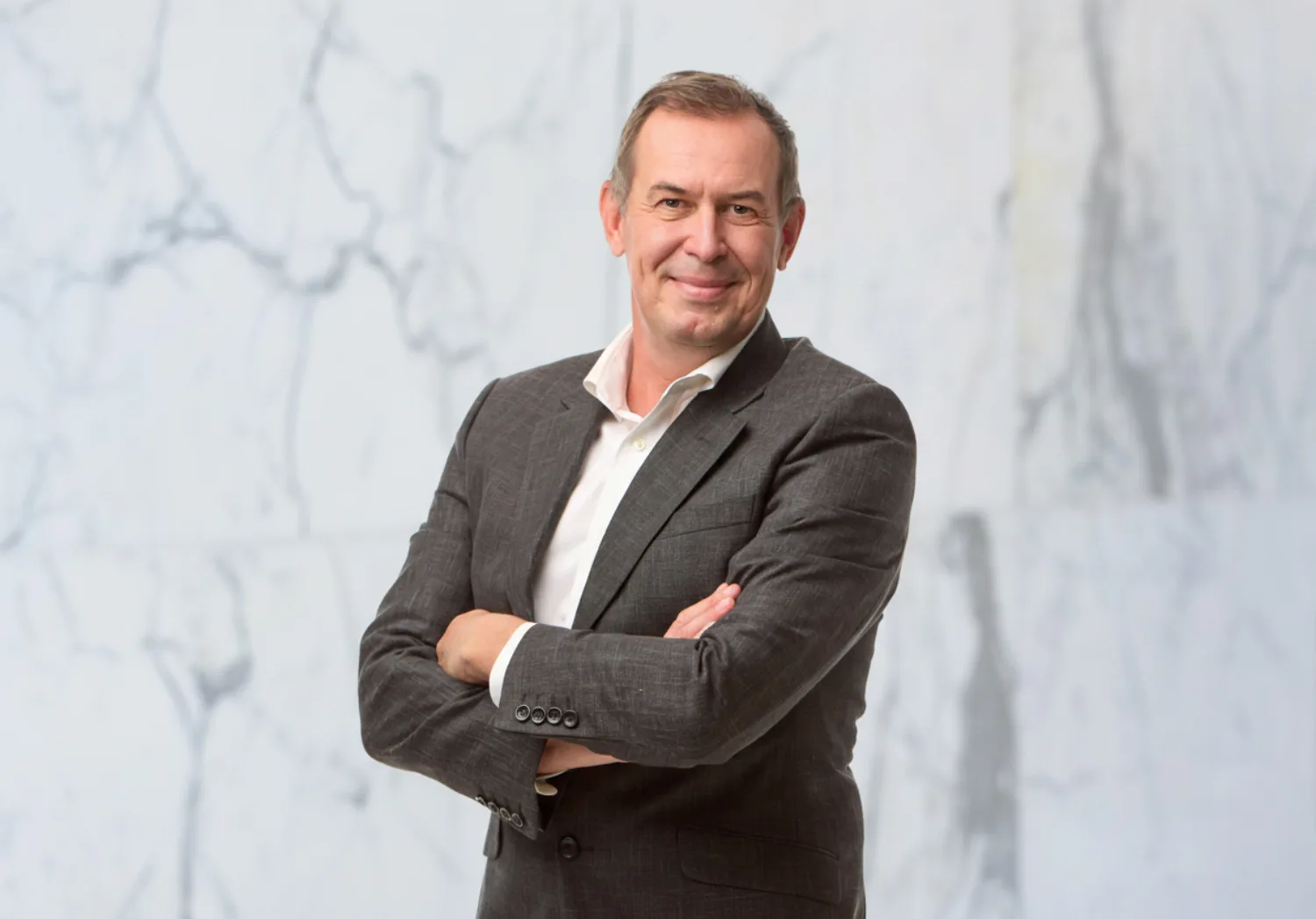 A person with short hair wearing a grey suit and white shirt stands with arms crossed in front of a white marble background, facing the camera.