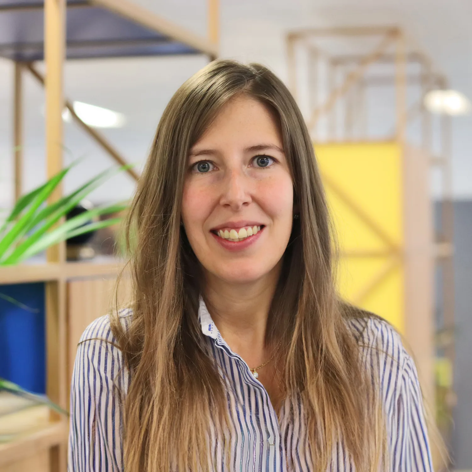 Head and shoulders portrait of Marta Hergueta Ortega wearing a stripy buttoned shirt with an office setting behind.