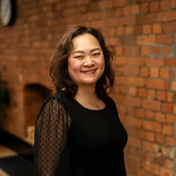 A person with shoulder‑length wavy hair stands indoors against a red‑brick wall. They wear a black top with sheer dotted sleeves and a small necklace. A plant and wall clock are visible in the background.