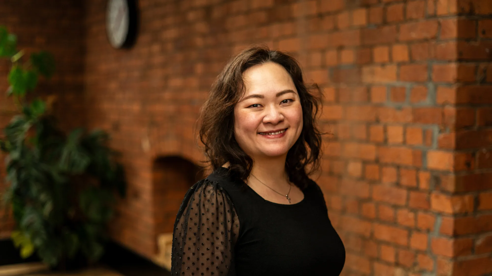 A person with shoulder‑length wavy hair stands indoors against a red‑brick wall. They wear a black top with sheer dotted sleeves and a small necklace. A plant and wall clock are visible in the background.