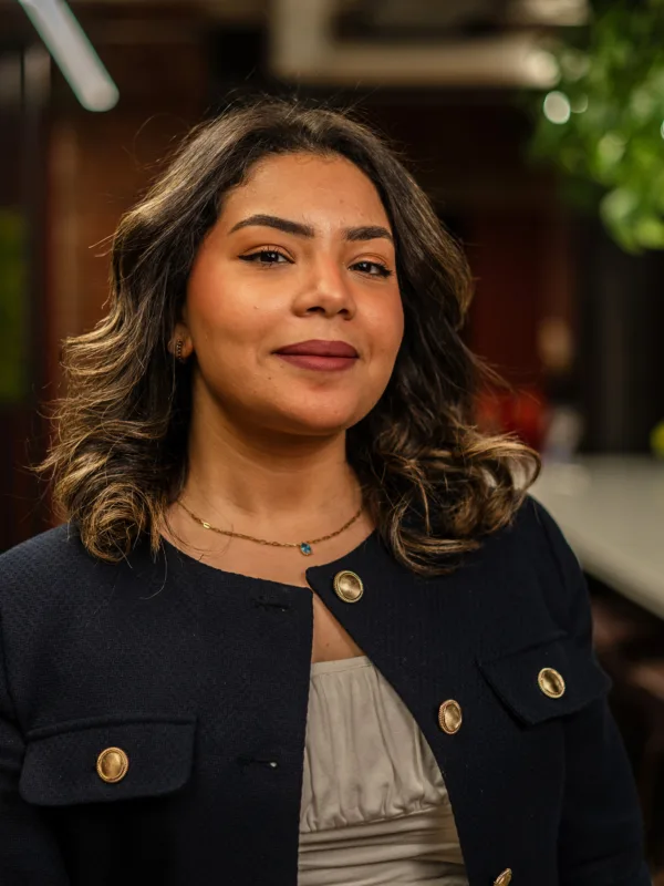 Dana in a dark blazer with gold buttons and a light top stands indoors. Background features a grey chair, green plants, and a long table in a modern lounge area.