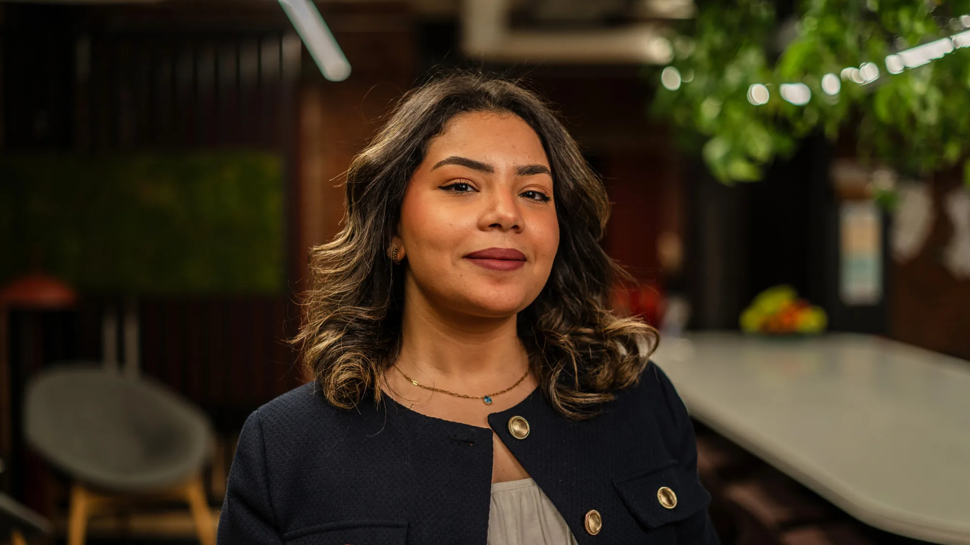 Dana in a dark blazer with gold buttons and a light top stands indoors. Background features a grey chair, green plants, and a long table in a modern lounge area.