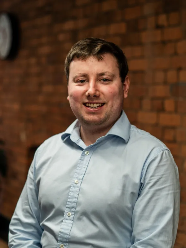 An individual in a light blue button‑up shirt sits indoors against a brick wall, with a clock on the wall and a large potted plant near a window in the background.