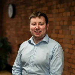 An individual in a light blue button‑up shirt sits indoors against a brick wall, with a clock on the wall and a large potted plant near a window in the background.